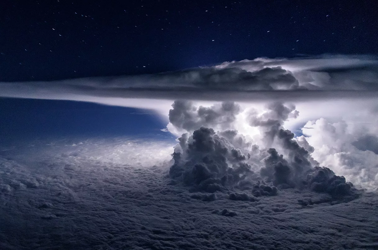 Uma colossal nuvem de tempestade vista de uma aeronave durante a noite. É possível ver o seu interior iluminado por raios, e estrelas no céu além.